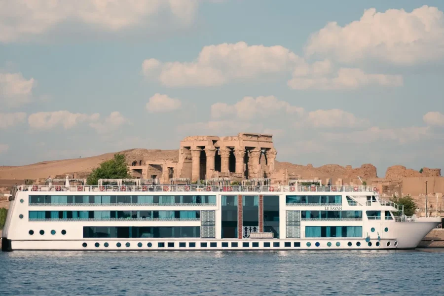 The MS Le Fayan Nile cruise ship on the Nile passing a stone temple standing on the bank behind it