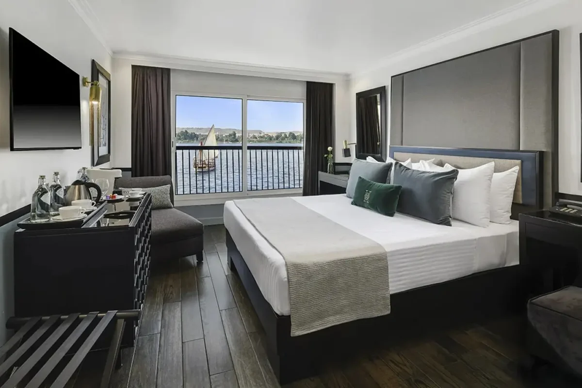 A cabin on the Movenpick MS Hamees Nile cruise with a wide bed and green cushions, a dark timber floor and a window looking onto a felucca on the Nile