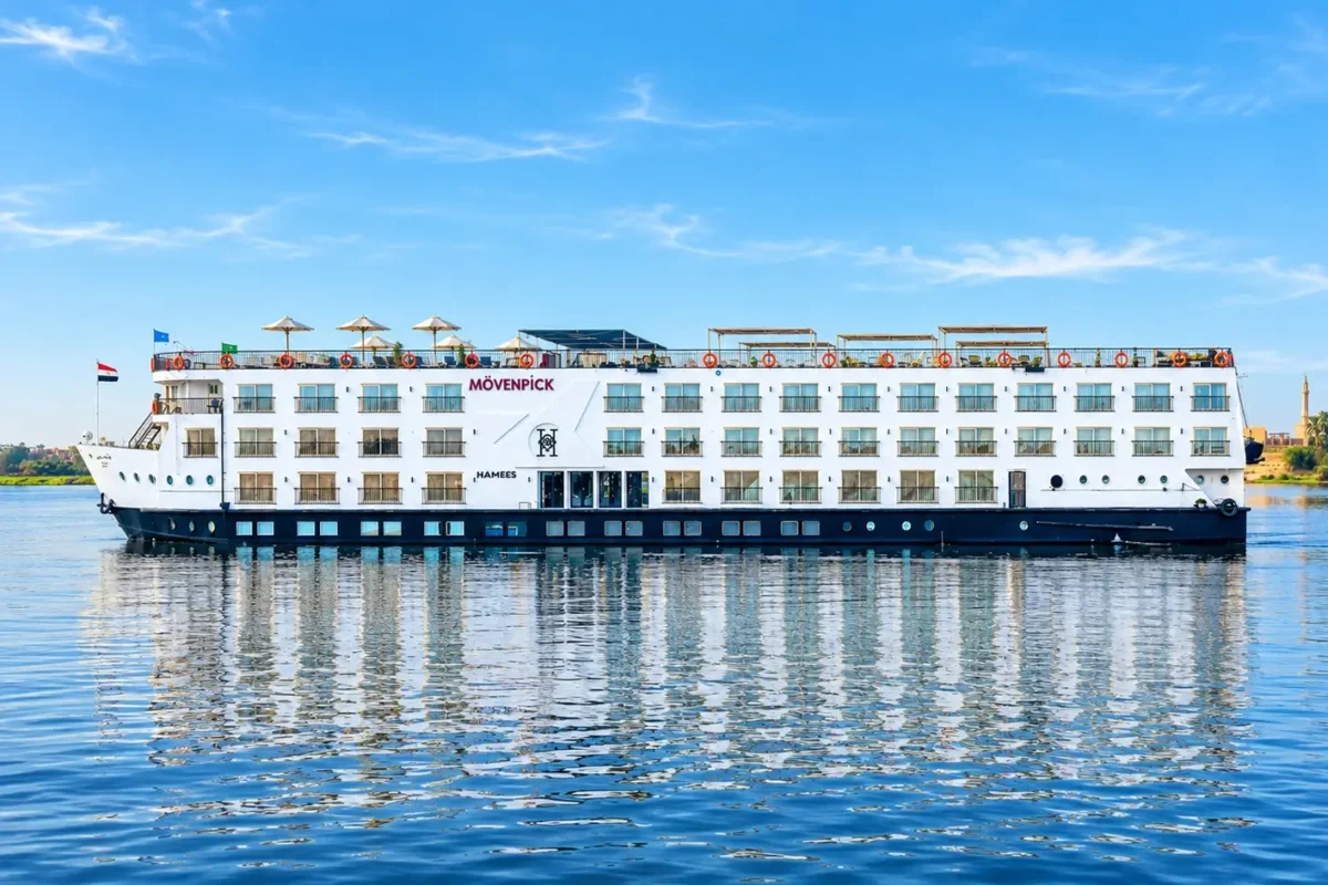 The Movenpick MS Hamees Nile cruise ship broadside on still water, its name along the upper deck and its reflection beneath it