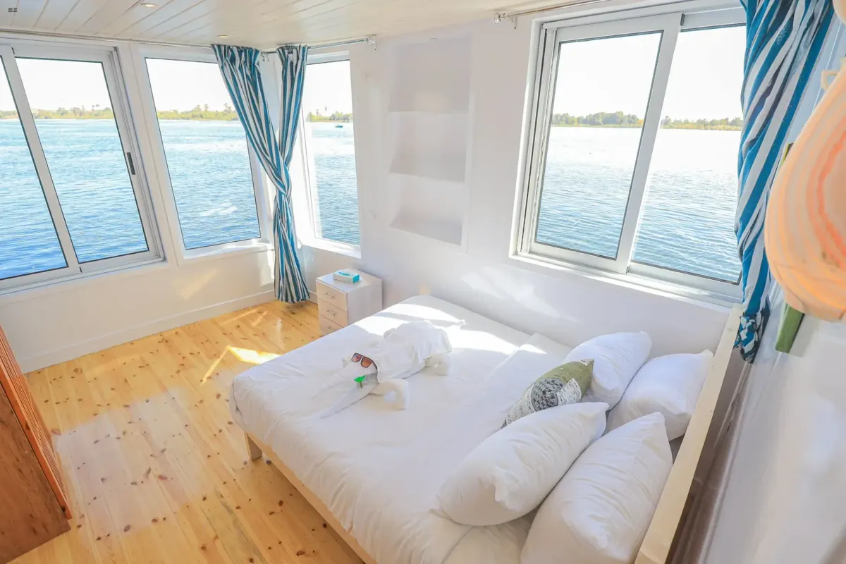 Un camarote de la dahabiya Nile Palace con cama doble y ventanas a dos lados sobre el río