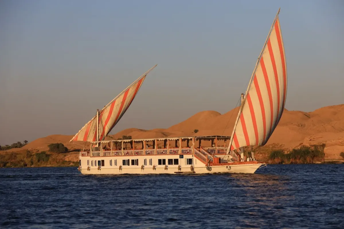 Die Dahabiya Nour El Din unter zwei gestreiften Segeln auf dem Nil, dahinter Wüstendünen am Westufer