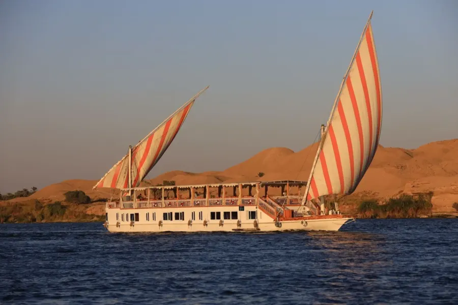 La dahabiya Nour El Din con dos velas rayadas en el Nilo, con dunas del desierto detrás de la orilla occidental