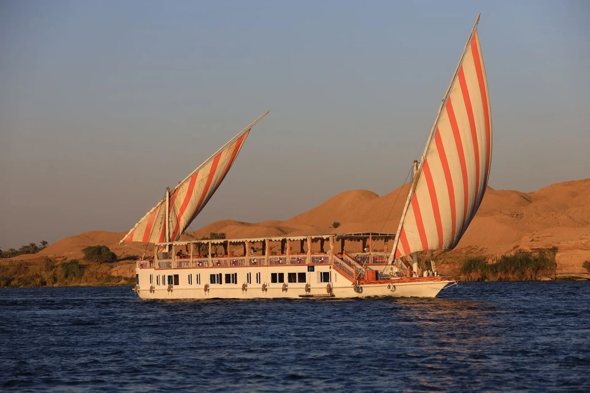 La dahabieh Nour El Din sous deux voiles rayées sur le Nil, avec des dunes désertiques derrière la rive ouest
