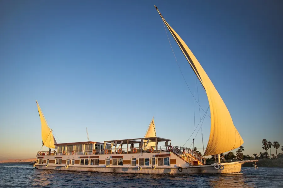 La dahabiya Princess Cleopatra bajo dos velas doradas al anochecer, con el casco blanco ribeteado en naranja
