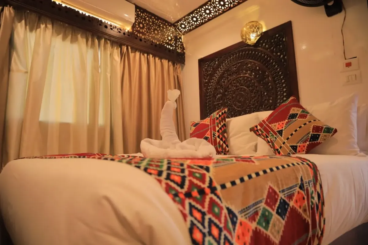 Un camarote de la dahabiya Queen Nefer con cama doble, cabecero tallado y textiles egipcios rojos