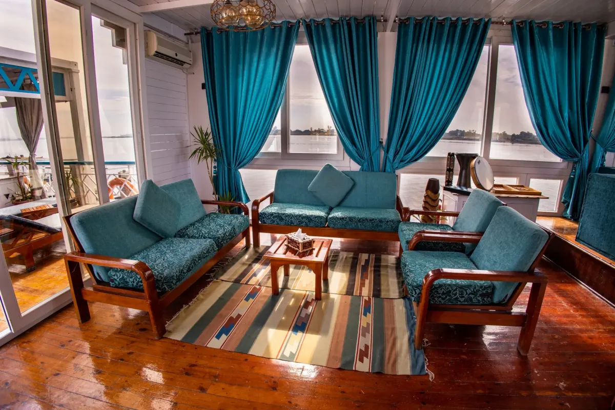 Der Salon der Dahabiya Royal Cleopatra, türkisfarbene Sofas und Sessel um einen niedrigen Tisch unter Fenstern zum Fluss