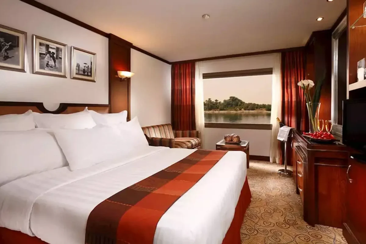 A double cabin on the Sonesta Nile Goddess Nile cruise with a red runner, framed prints and a window onto the riverbank