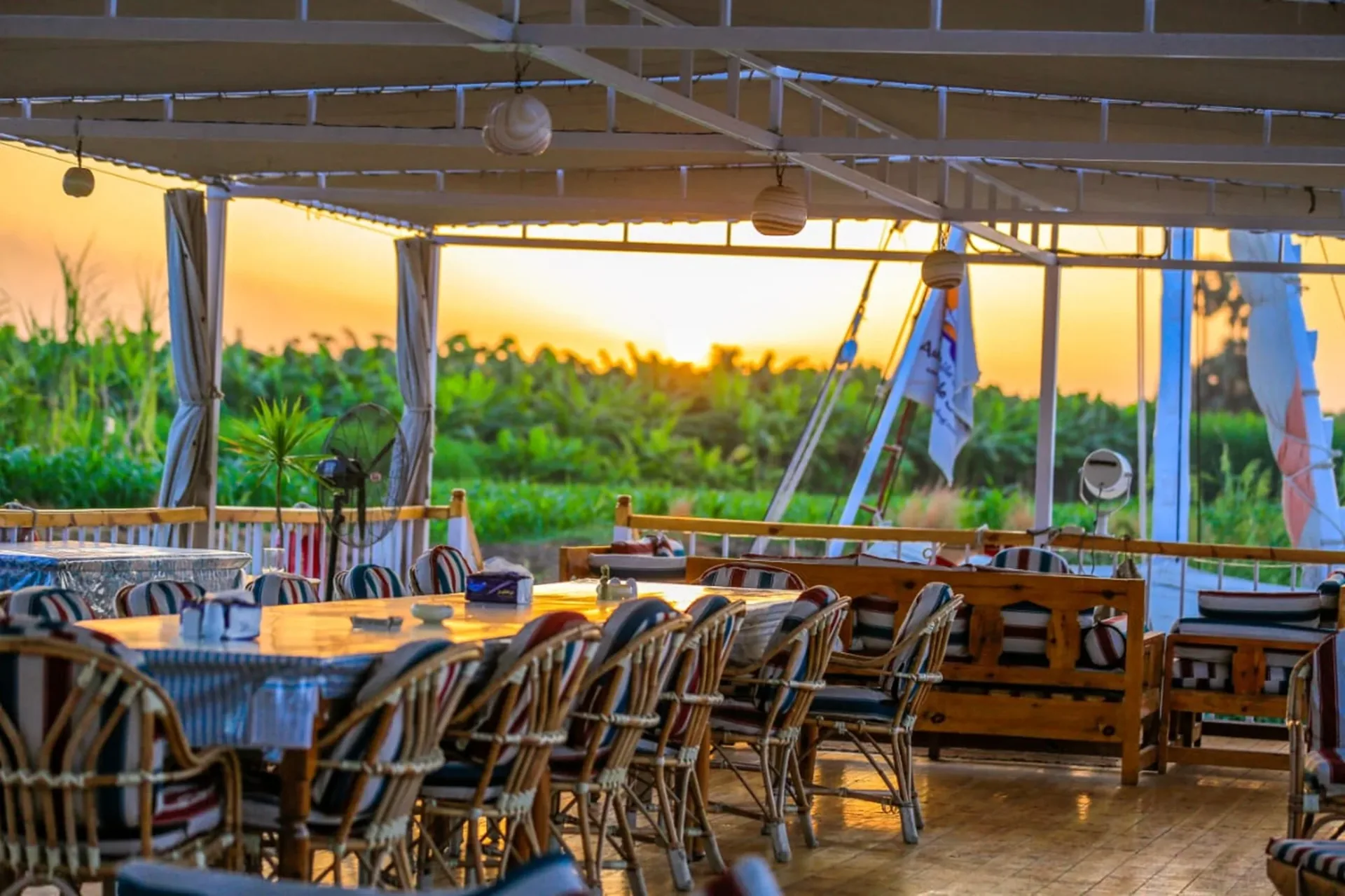 Des tables pour les repas sur le pont ombragé de la dahabieh Three Pyramids au coucher du soleil, avec des palmiers sur la rive
