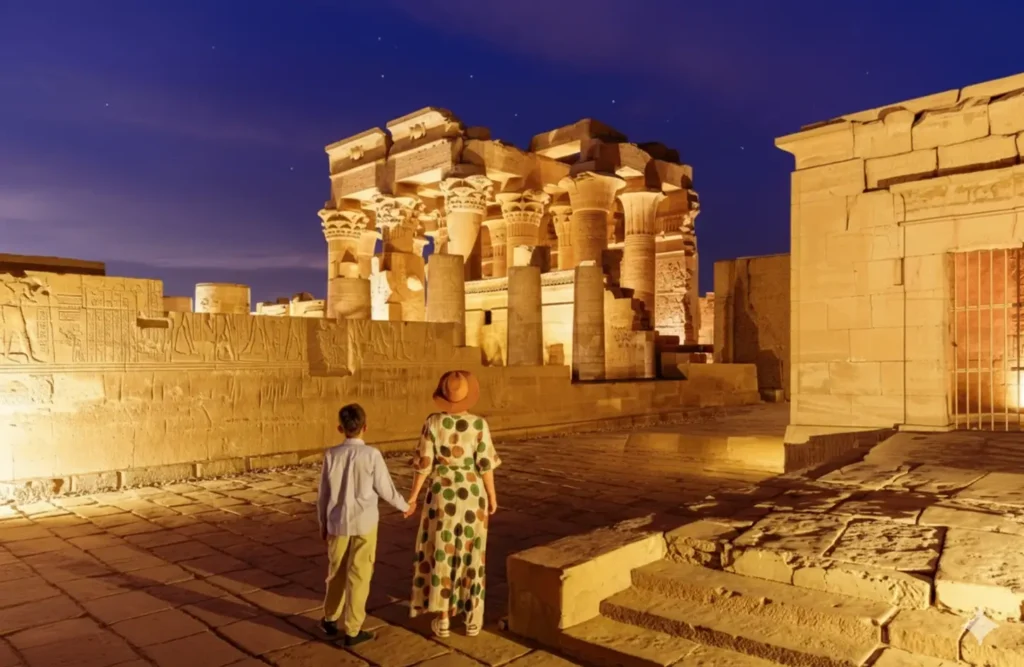 A couple walking towards the floodlit Temple of Kom Ombo under a deep blue evening sky