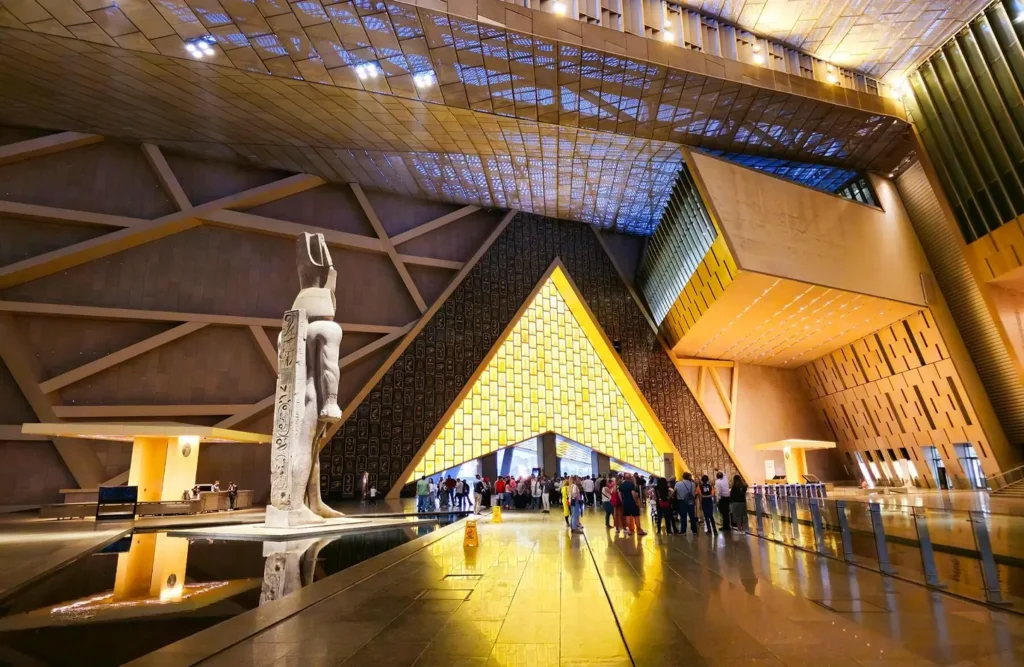 The colossal statue of Ramses II in the grand hall of the Grand Egyptian Museum, visitors queuing beneath the glazed roof
