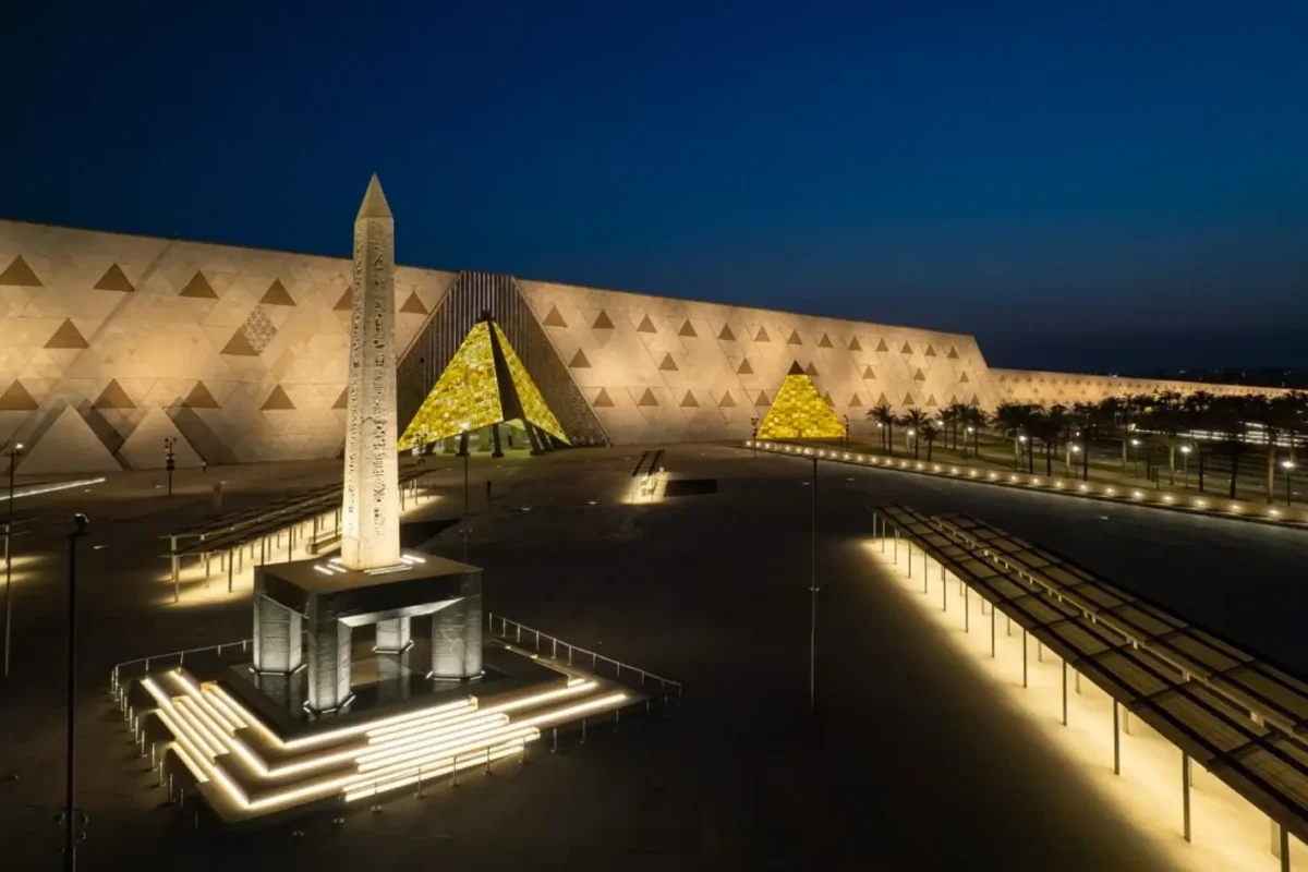 The hanging obelisk lit in the forecourt of the Grand Egyptian Museum at night, the museum's triangular façade glowing behind