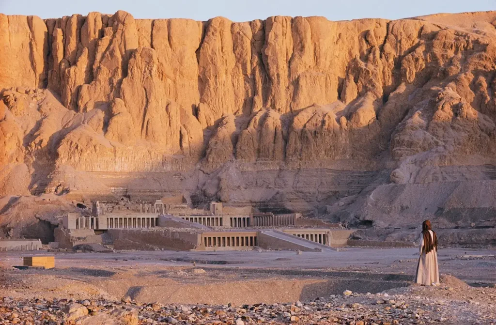The Temple of Hatshepsut at the foot of its cliffs in early light, a lone figure standing on the desert ground in front