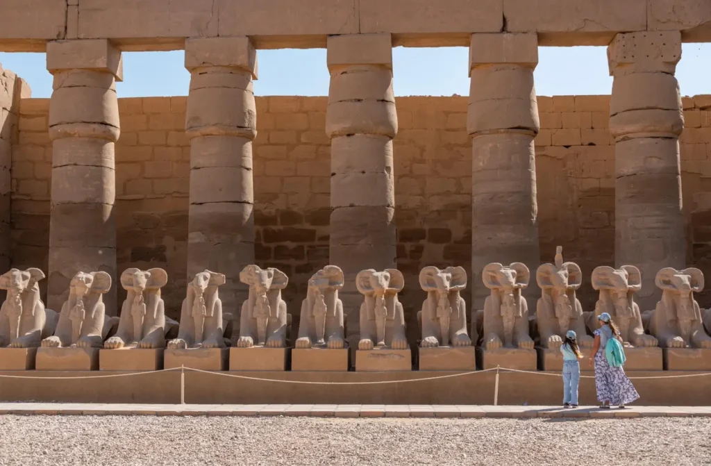 The row of ram-headed sphinxes along the forecourt at Karnak Temple, two visitors walking past them