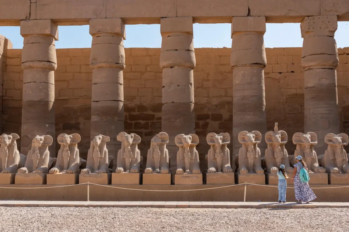 The row of ram-headed sphinxes along the forecourt at Karnak Temple, two visitors walking past them