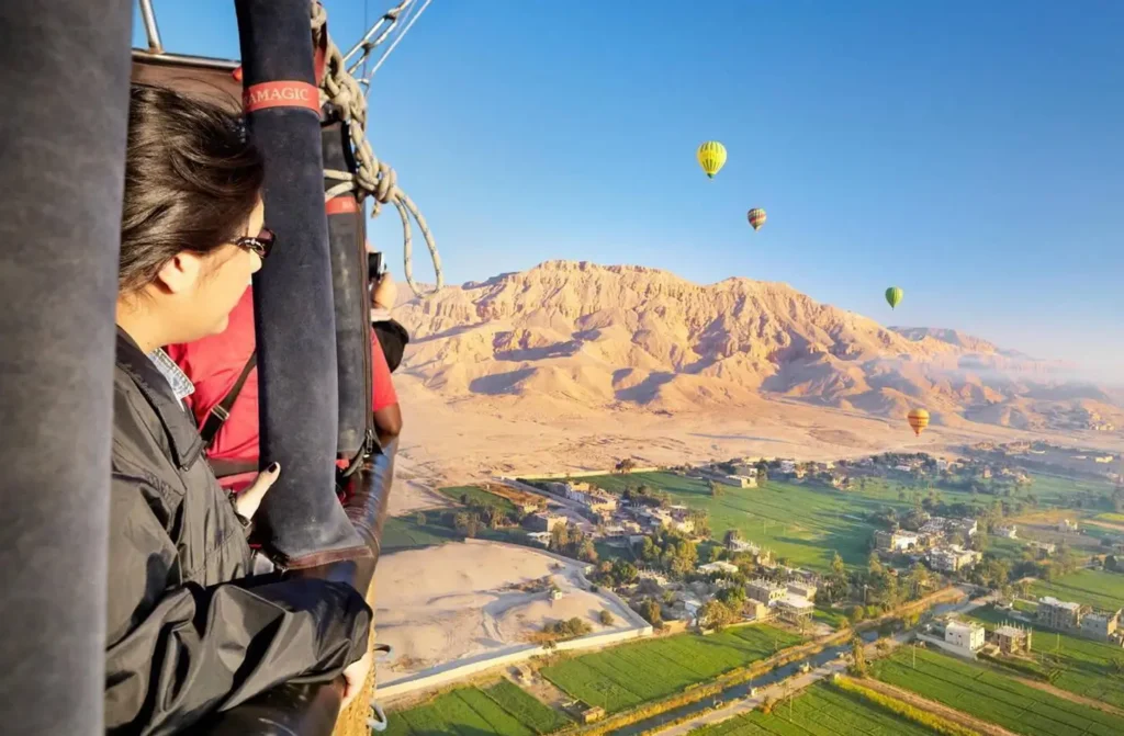 A passenger looking out from a balloon basket over Luxor, other balloons rising above the desert cliffs