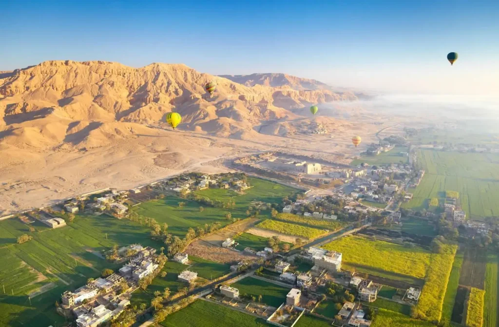 The Luxor west bank from a balloon at dawn, cultivated fields meeting the pale Theban cliffs