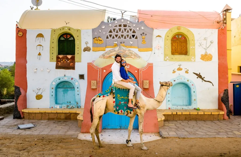 A man on a camel in front of a Nubian house painted with bright patterns and arched doorways