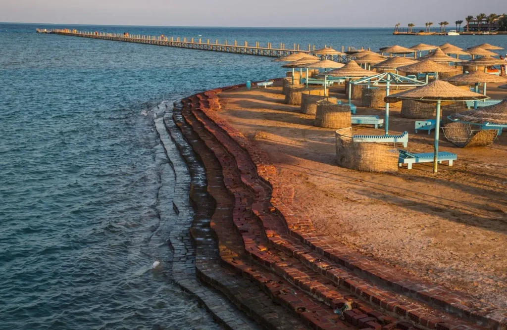 Straw parasols and sun loungers lining a Red Sea beach seen from above, a jetty running out into the water