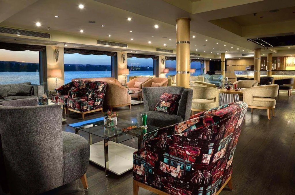 The lounge on the MS Acamar Nile cruise, patterned armchairs around glass tables in front of floor-to-ceiling river windows