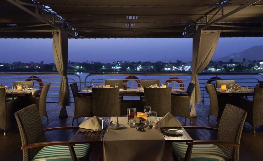 Open-air dining tables under the shaded deck of the MS Acamar Nile cruise at twilight, the lights of the far bank across the water