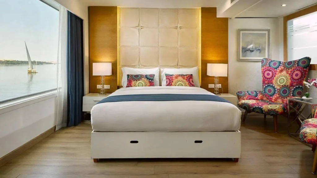 A suite on the MS Acamar Nile cruise with a king bed, upholstered headboard and a picture window framing a felucca on the river