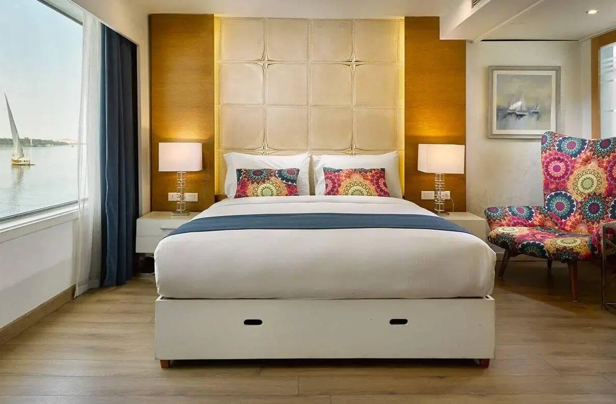 A suite on the MS Acamar Nile cruise with a king bed, upholstered headboard and a picture window framing a felucca on the river