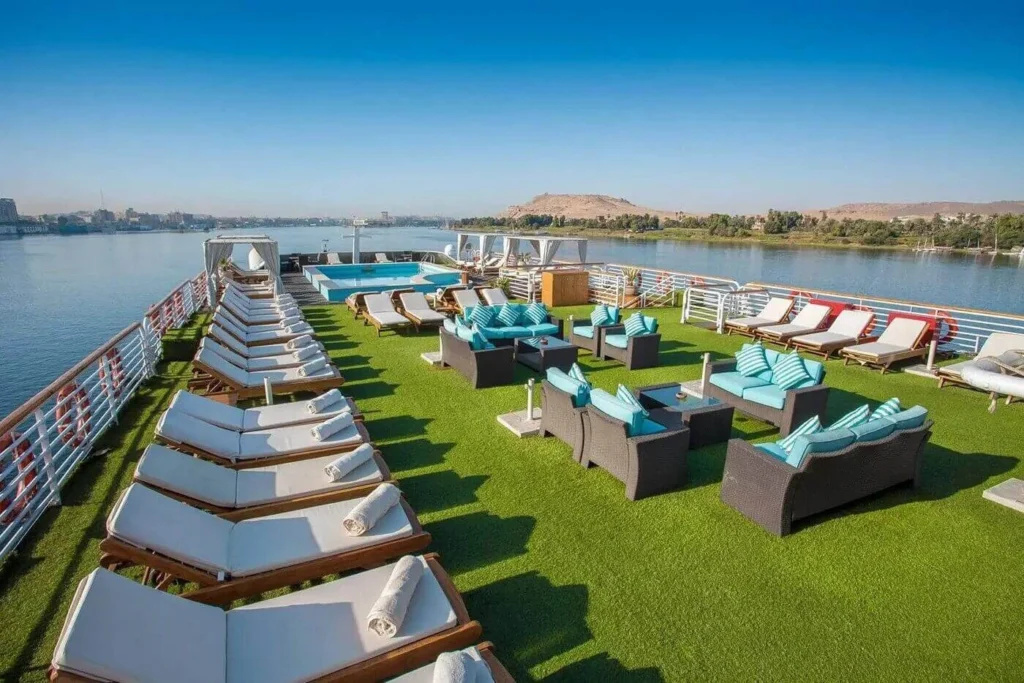The sun deck of the MS Acamar Nile cruise with rows of loungers and turquoise daybeds on artificial lawn, the west bank hills beyond
