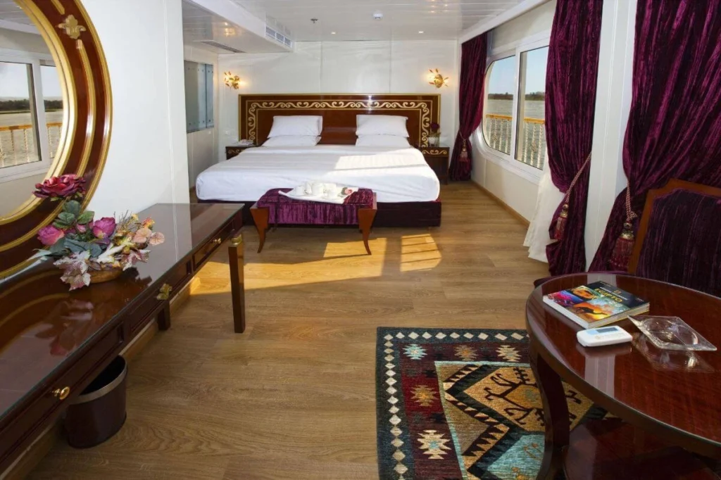 A suite on the Al Hambra Nile cruise with a carved bedhead, a long writing desk, a gilt mirror and burgundy drapes at the river window