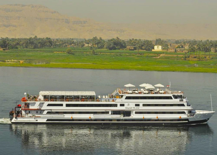 The MS Alexander the Great Nile cruise ship moored beside cultivated fields, the desert escarpment pale behind them