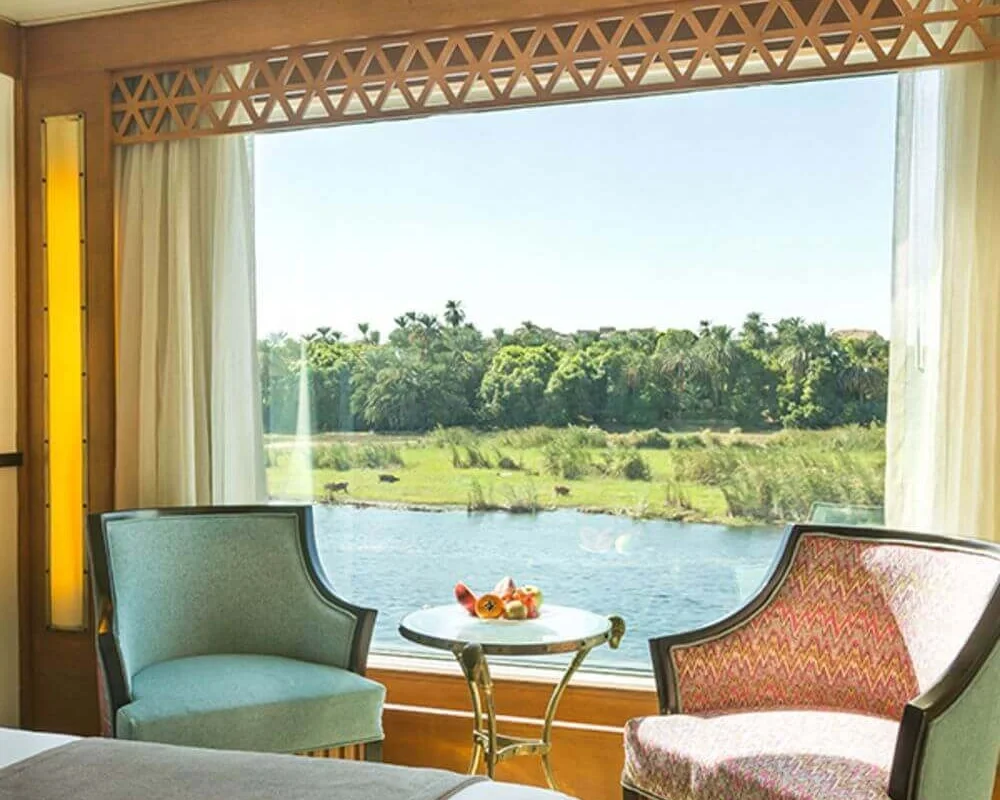 Two armchairs and a small table set with fruit at a cabin window on the Alyssa Nile cruise, looking across the river to a green bank