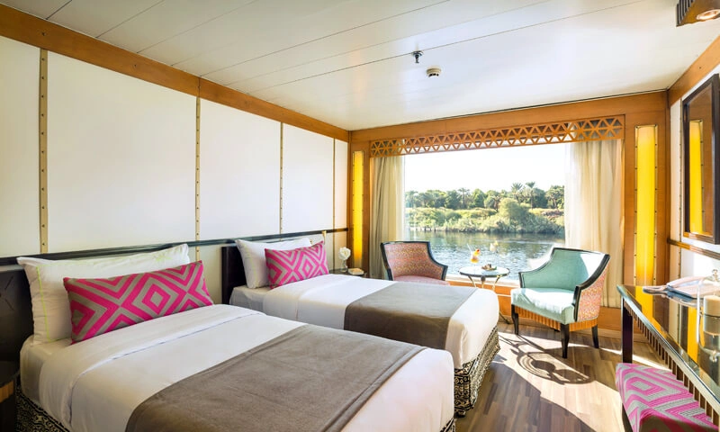 A twin-bed cabin on the Alyssa Nile cruise with pink cushions, a seating corner and a wide window framing the riverbank