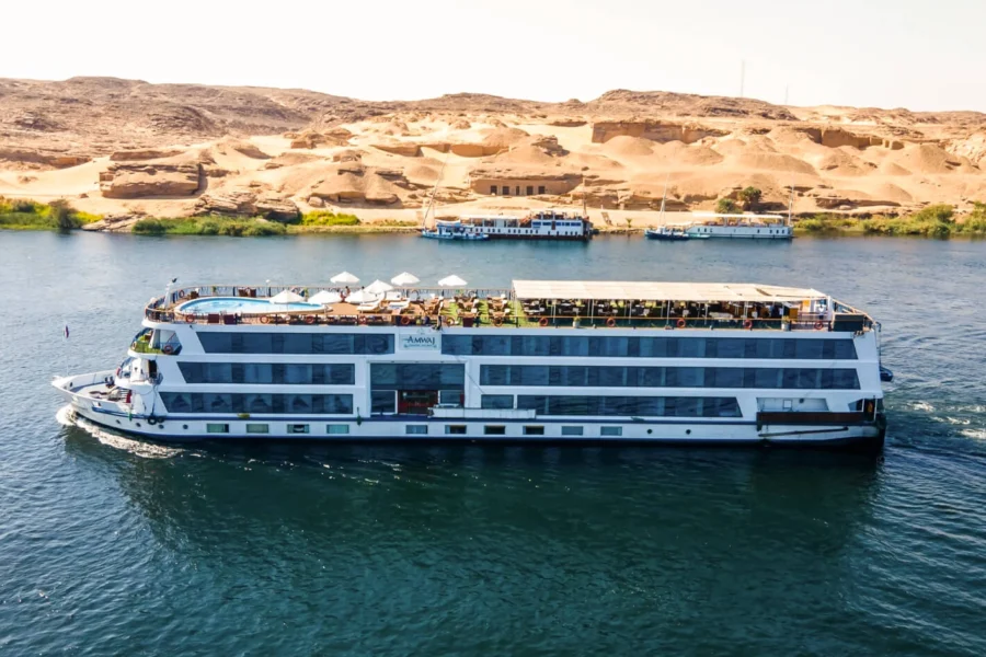 The MS Amwaj Living Stone Nile cruise ship on the Nile below sand dunes and rock, other cruisers moored along the far bank
