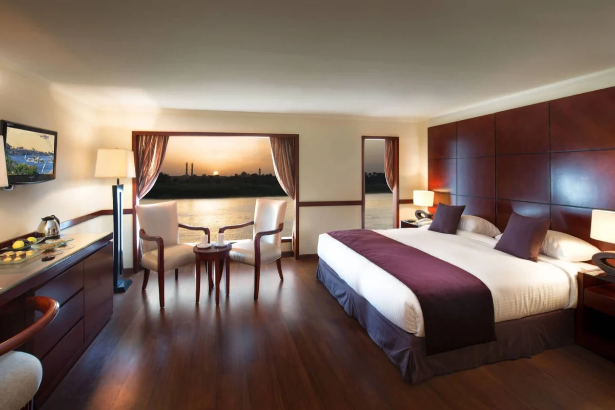 A suite on the MS Amwaj Living Stone Nile cruise with a dark wood floor, a king bed, a writing desk and two chairs at the window