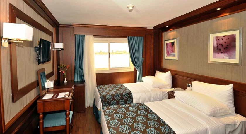 A twin-bed cabin on the Farida Nile cruise with a patterned runner, a writing desk and turquoise curtains at the river window