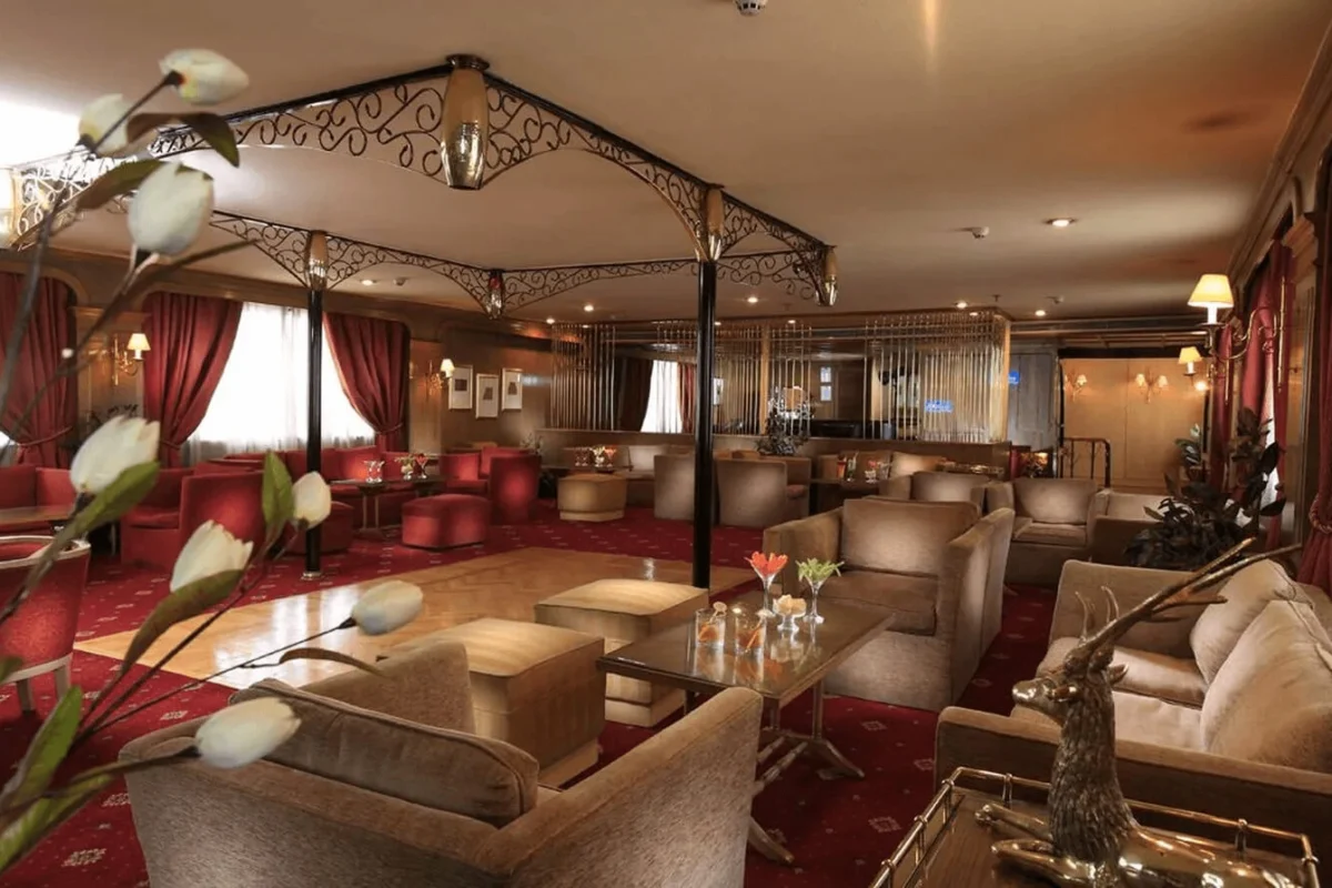 Le salon principal de la croisière Iberotel Amara sur le Nil, avec canapés et fauteuils sur un tapis rouge sous une verrière en fer forgé