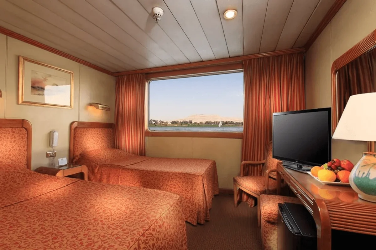 A twin-bed cabin on the Iberotel Amara Nile cruise in warm terracotta tones, a television and fruit bowl on the console beneath the river window