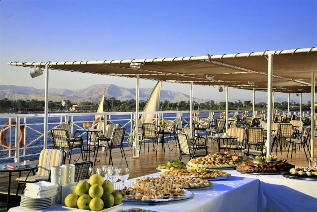 A buffet table of pastries and fruit set out on the shaded deck of the Iberotel Crown Emperor Nile cruise, dining tables beyond