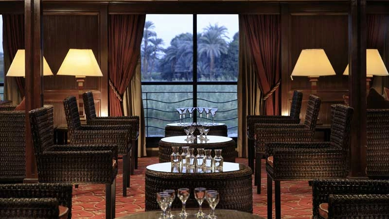 Wicker armchairs and table lamps beside the tall lounge window of the Iberotel Crown Empress Nile cruise, palms visible outside