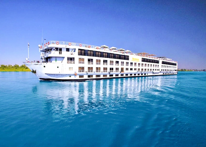 The Iberotel Crown Empress Nile cruise ship on bright blue water, seen from the bow with a green bank behind