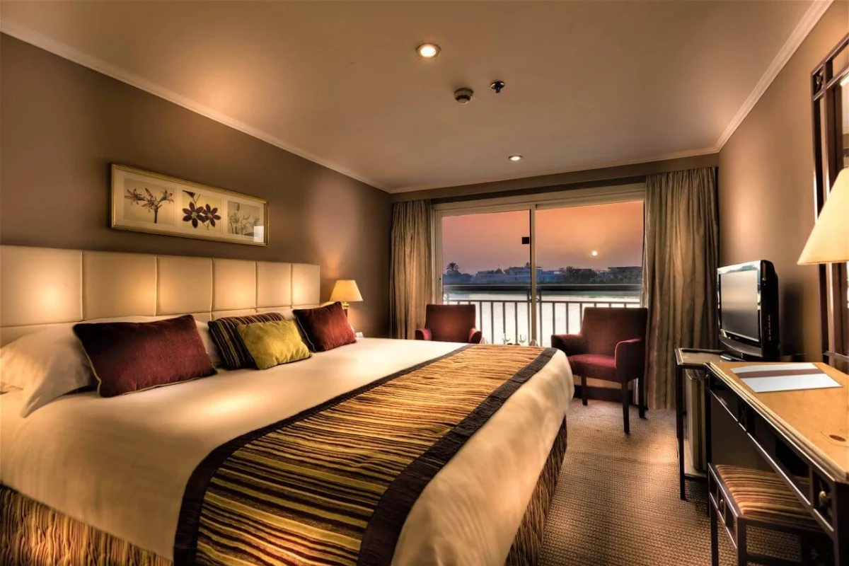 A suite on the Jaz Crown Jewel Nile cruise with a wide bed, a writing desk and a glass door onto a private balcony at dusk