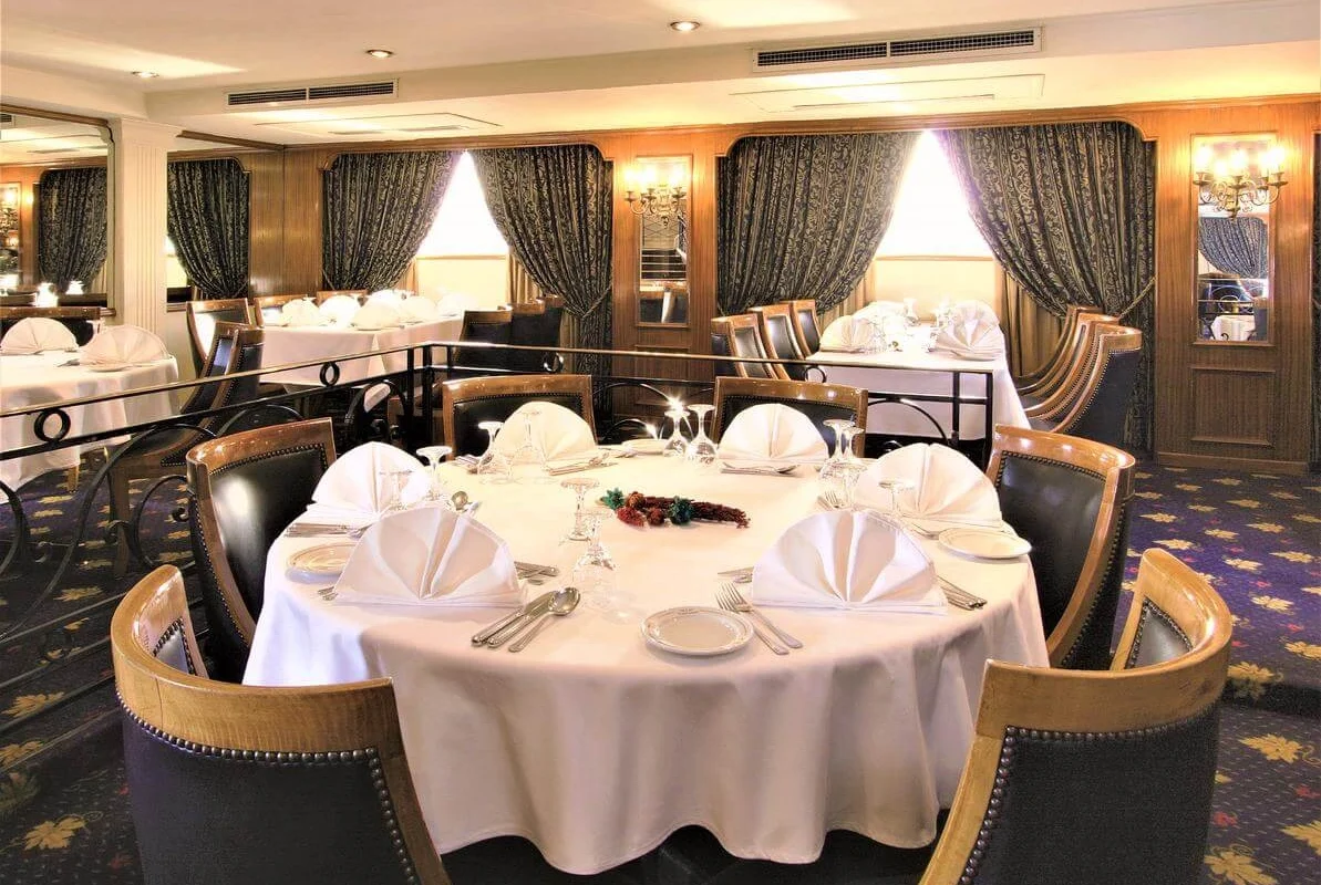 A round table laid with white linen and fan-folded napkins in the Jaz Crown Prince Nile cruise restaurant, curtained windows behind