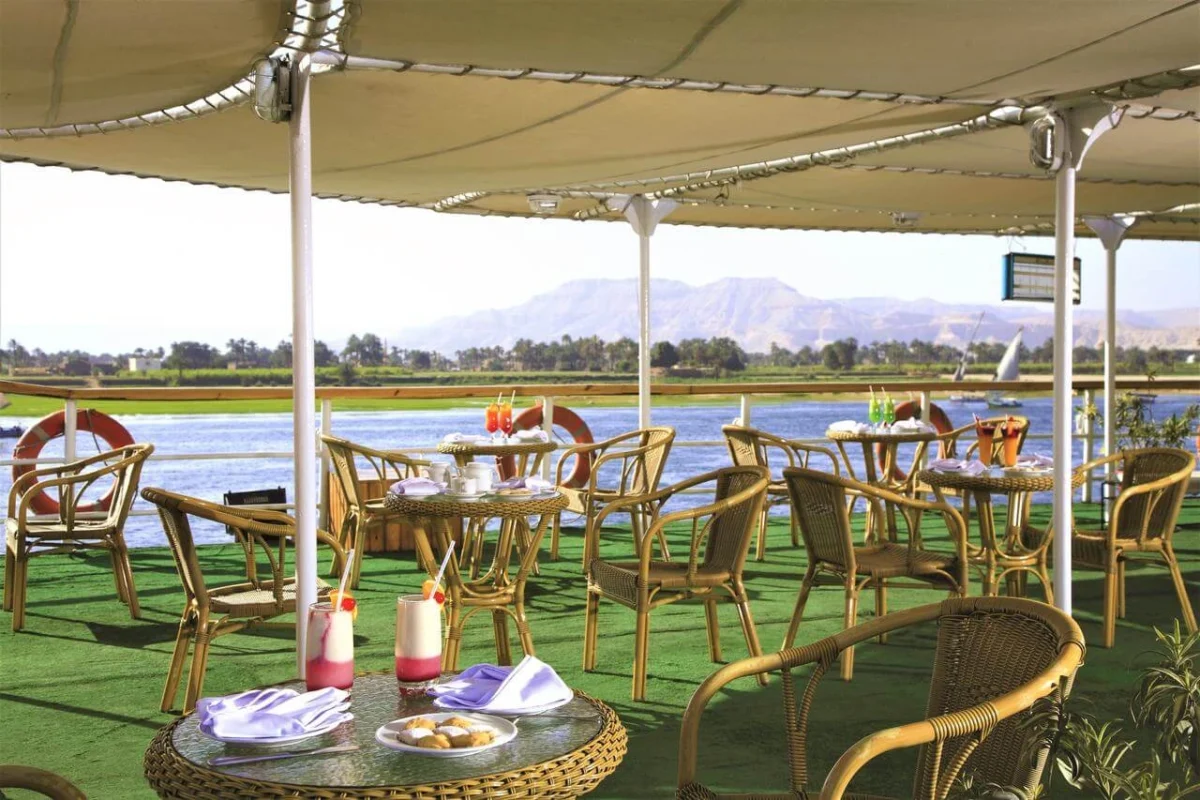 Cane chairs and tables under the deck canopy of the Jaz Crown Prince Nile cruise, the Theban hills across the water