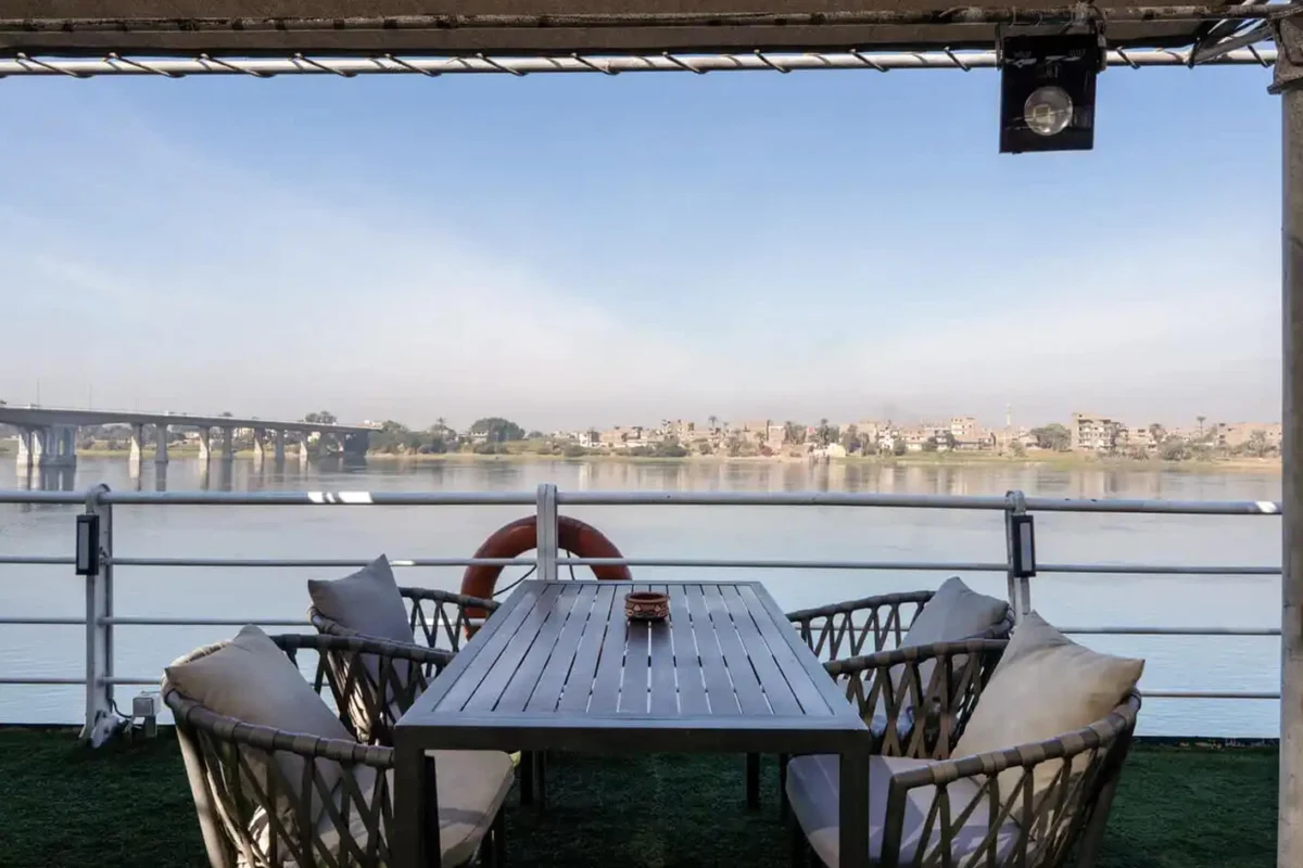 A table and cushioned chairs at the rail on the Magic I Nile cruise deck, the far bank across open water