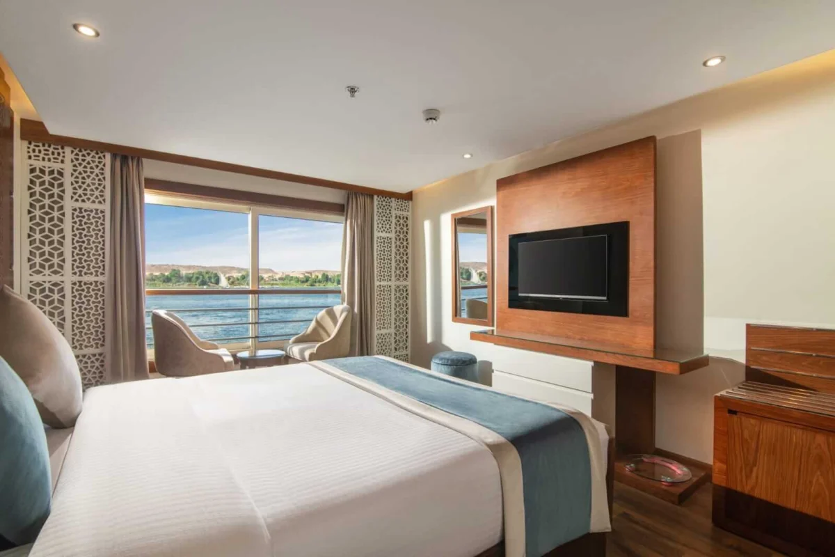 A double cabin on the Nile Capital Nile cruise with teal cushions, a television and armchairs at the window onto the river
