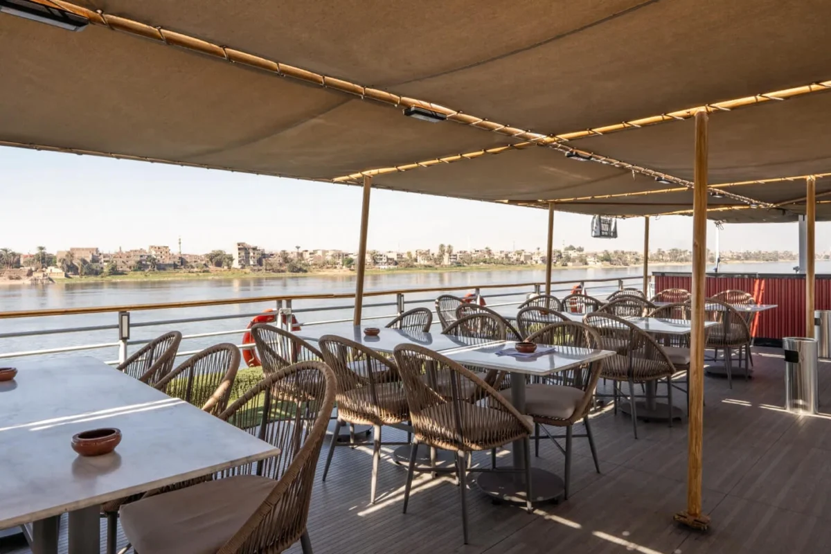 Woven chairs and tables under a canvas awning on the Nile Excellence Nile cruise deck, a town along the far bank