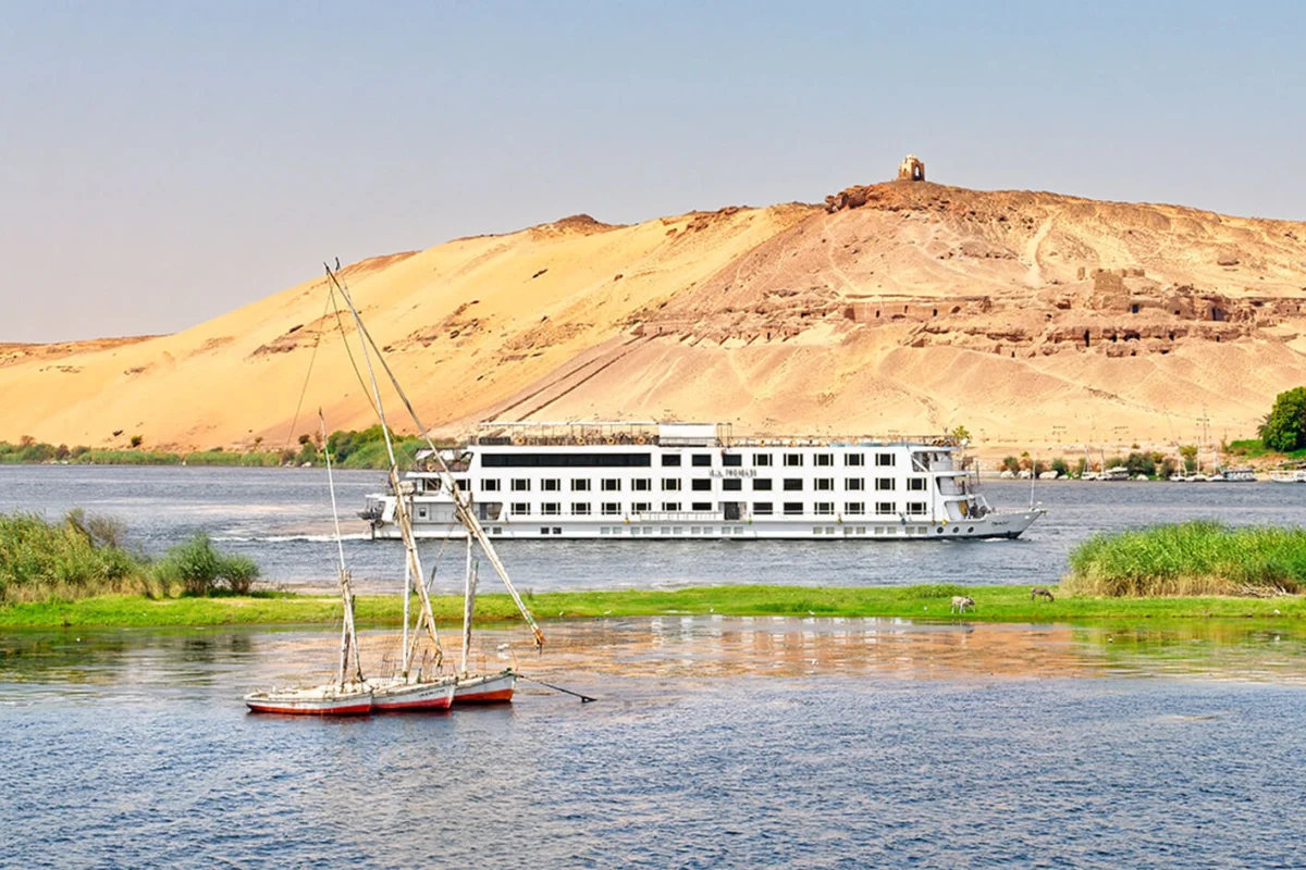 The Nile Premium Nile cruise ship moored below a sand hill topped by a ruined tomb, a felucca in the foreground