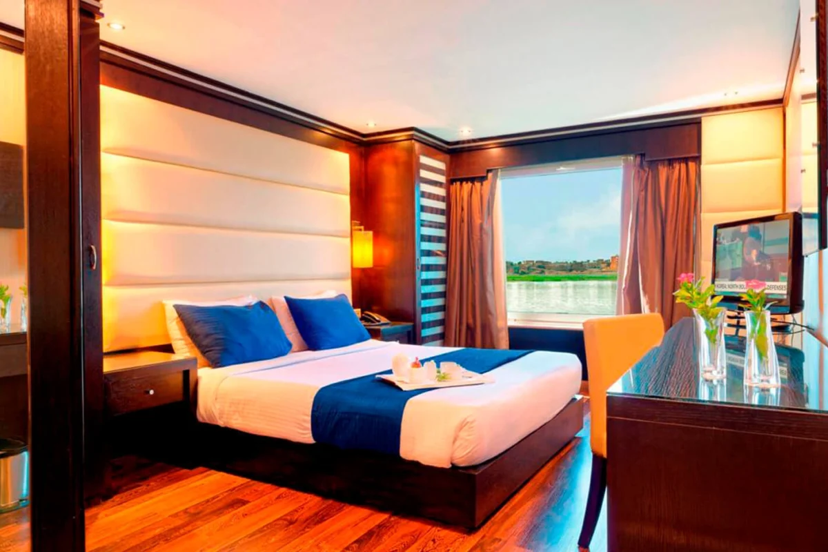 Eine Suite auf der Nilkreuzfahrt Nile Premium mit Himmelbett, blauen Kissen und einem Fenster zu grünen Feldern