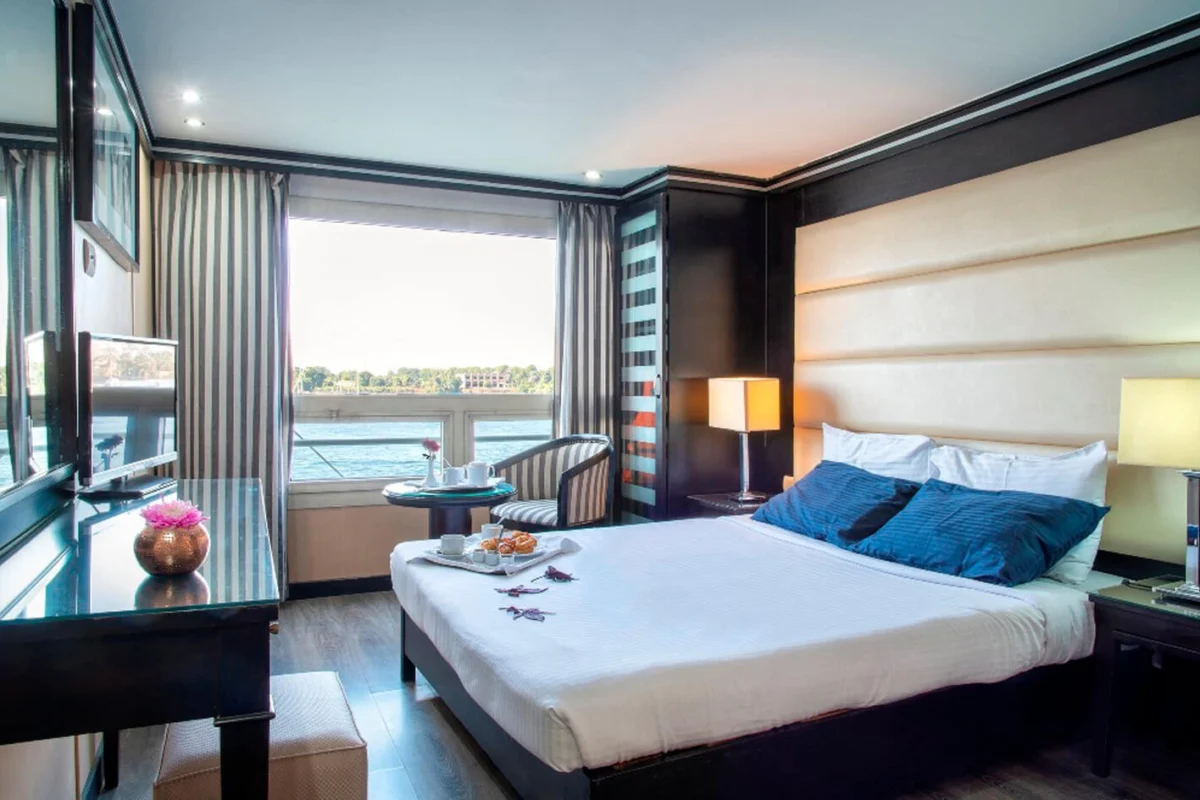 A suite on the Nile Style Nile cruise with a padded headboard wall, blue cushions and a glass door onto the balcony