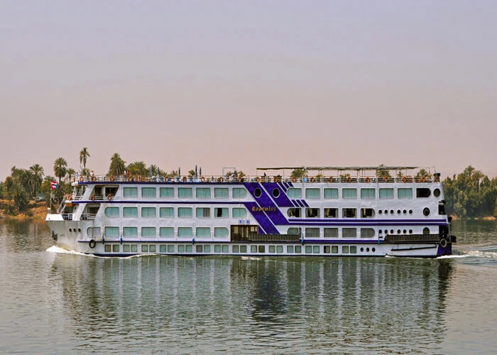 The Radamis I Nile cruise ship on the Nile with its blue chevron marking, palms along the bank behind