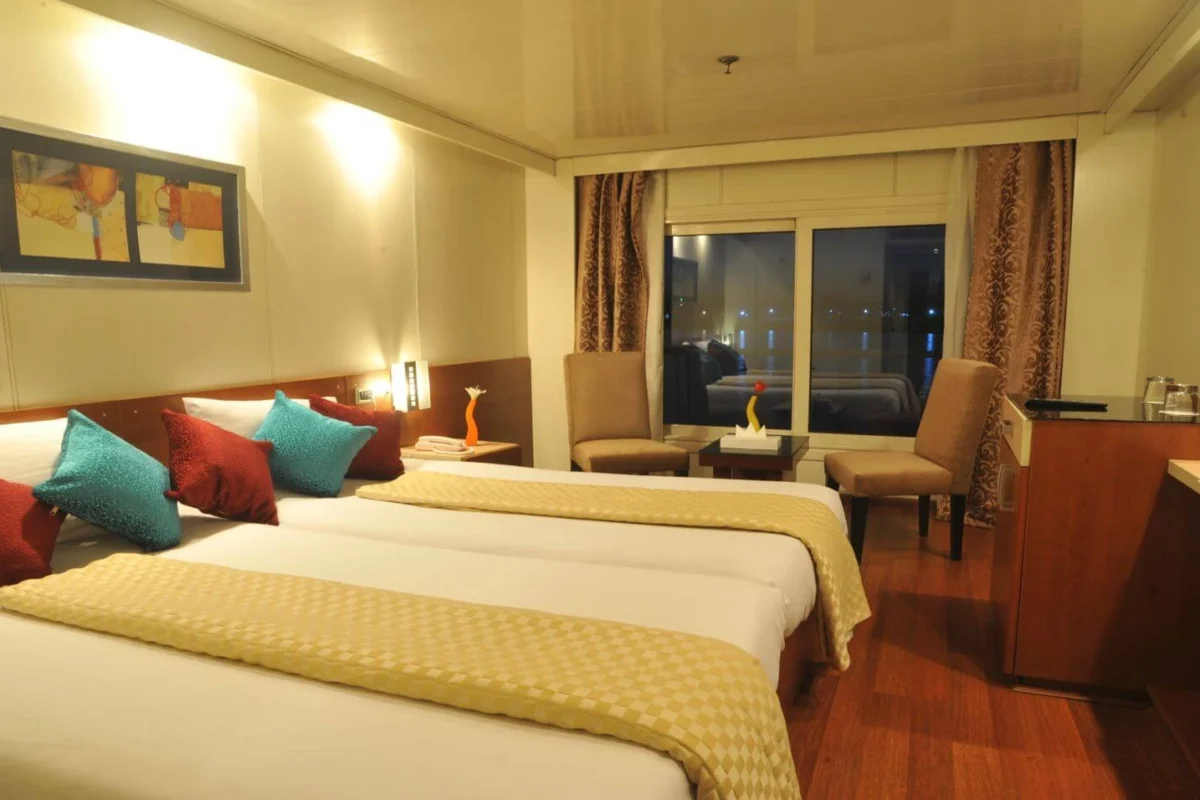 A twin-bed cabin on the Royal Esadora Nile cruise with gold covers and teal cushions, a desk and armchair by the window